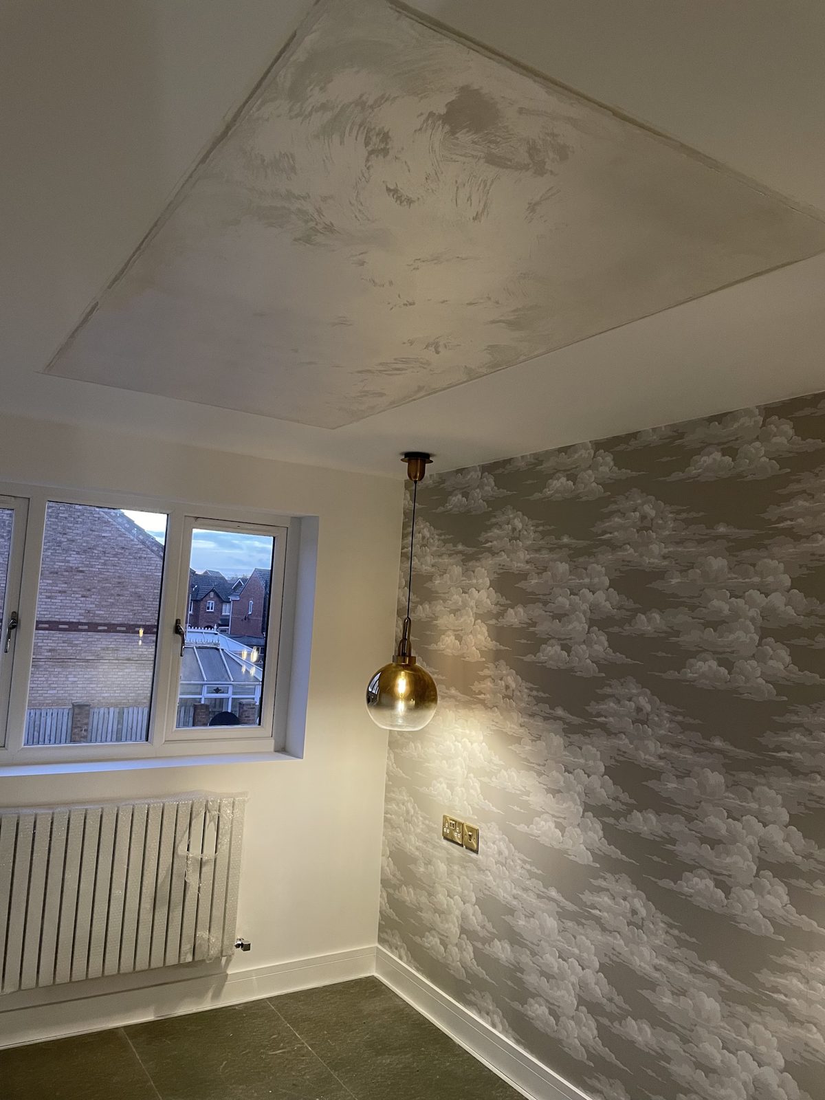 A bedroom with a brass pendant light, cloud wallpaper and a softly lit ceiling recess