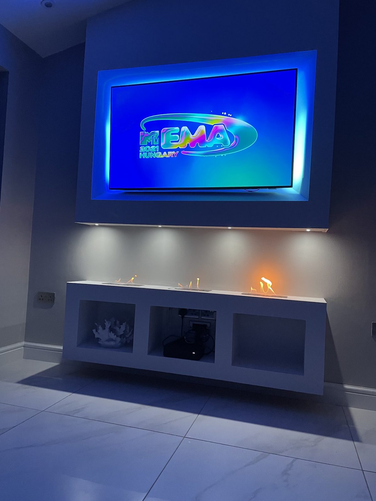 A media wall with blue LED backlighting around the television, downlights and a lit fireplace below