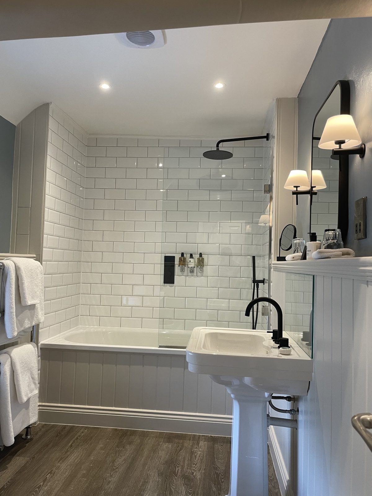 A finished bathroom with downlights, swan-neck wall lights over the basin and white metro tiles